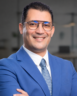 Ein Mann in einem blauen Anzug mit Brille lächelt, während er mit verschränkten Armen vor einem modernen Bürohintergrund steht.