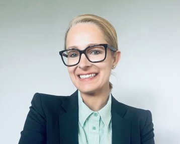 Eine Frau mit blonden Haaren trägt eine schwarze Blazerjacke und ein gestreiftes Hemd. Sie lächelt und hat die Arme verschränkt.
