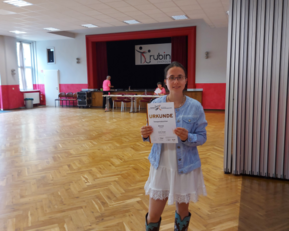 Ein Mädchen hält eine Urkunde in der Hand, steht in einem großen Raum mit Holzboden und einem roten Vorhang im Hintergrund.