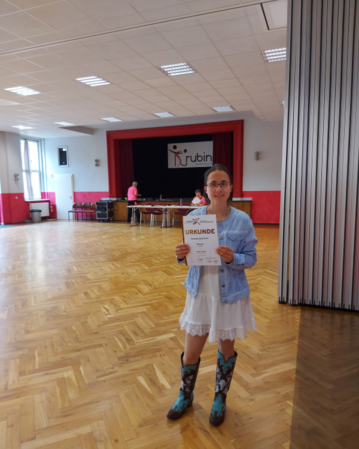 Ein Mädchen hält eine Urkunde in der Hand, steht in einem großen Raum mit Holzboden und einem roten Vorhang im Hintergrund.