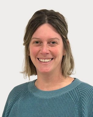 Eine lächelnde Person mit mittellangen, blonden Haaren trägt einen blauen Pullover. Der Hintergrund ist neutral und hell.