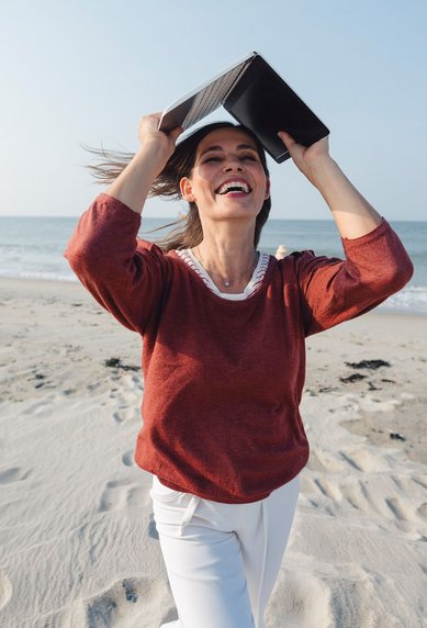 Eine Frau läuft am Strand und hält ein Buch über ihrem Kopf, während sie lächelt und die Meereswellen im Hintergrund sichtbar sind.