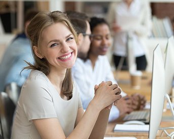 Eine lächelnde Frau sitzt an einem Schreibtisch mit einem Computer, umgeben von anderen Personen in einem modernen Büro. Sie trägt ein helles Oberteil und hat lange, lockige Haare.