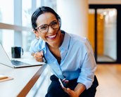 Eine lächelnde Frau mit Brille sitzt an einem Tisch mit einem Laptop und einer Tasse, während sie ein Smartphone in der Hand hält.