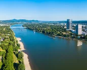 Luftaufnahme des Rheins mit grünen Uferbereichen und modernen Gebäuden im Hintergrund. Die Landschaft zeigt eine Mischung aus Natur und urbaner Entwicklung.