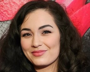 Eine Frau mit lockigem, dunklem Haar und einem lila Oberteil lächelt in die Kamera, vor einem bunten Hintergrund.