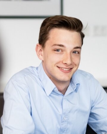 Ein junger Mann mit braunen Haaren trägt ein hellblaues Hemd und lächelt freundlich, während er an einem Tisch sitzt.