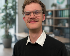 Junger Mann mit Brille und Schnurrbart, trägt einen schwarzen Pullover und ein weißes Hemd, steht in einem modernen Büro mit Pflanzen im Hintergrund.