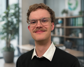 Junger Mann mit Brille und Schnurrbart, trägt einen schwarzen Pullover und ein weißes Hemd, steht in einem modernen Büro mit Pflanzen im Hintergrund.
