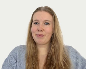 Eine Frau mit langen, blonden Haaren trägt einen grauen Pullover und schaut direkt in die Kamera. Der Hintergrund ist neutral.