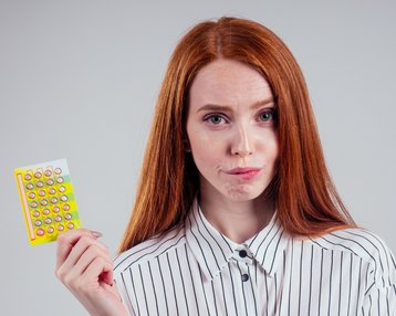 Eine Frau mit langen roten Haaren hält eine Packung Antibabypillen in der Hand und schaut neutral in die Kamera. Sie trägt ein gestreiftes Hemd.