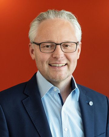 Ein Mann mit Brille und grauen Haaren trägt einen dunklen Anzug und ein hellblaues Hemd. Er lächelt vor einem farbigen Hintergrund in Orange und Blau.