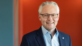 Ein Mann mit Brille und grauen Haaren trägt einen dunklen Anzug und ein hellblaues Hemd. Er lächelt vor einem farbigen Hintergrund in Orange und Blau.