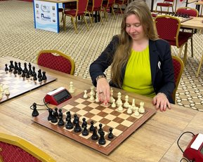 Eine Schachspielerin mit langen Haaren in einem schwarzen Blazer und gelbem Oberteil zieht eine Figur auf einem Schachbrett.