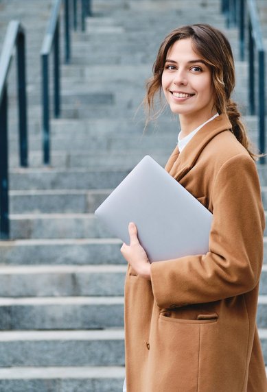 Junge Frau mit braunem Mantel steht an einer Treppe und hält einen Laptop in der Hand. Sie lächelt in die Kamera.
