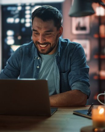 Ein Mann sitzt an einem Tisch und lächelt, während er auf einen Laptop schaut. Neben ihm stehen eine Tasse und eine Kerze. Im Hintergrund sind Bücherregale und Fenster mit Stadtansicht zu sehen.