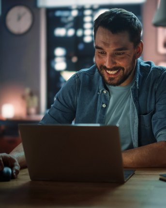 Gesundheitspsychologie berufsbegleitend studieren Ein Mann sitzt an einem Tisch und lächelt, während er auf einen Laptop schaut. Neben ihm stehen eine Tasse und eine Kerze. Im Hintergrund sind Bücherregale und Fenster mit Stadtansicht zu sehen.