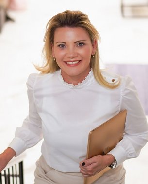 Eine Frau mit blonden Haaren trägt eine weiße Bluse und einen beigen Rock. Sie hält eine Mappe in der Hand und lächelt, während sie eine Treppe hinaufgeht.