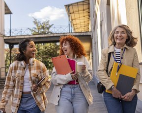 Drei Frauen gehen lächelnd auf einem Gehweg, tragen Ordner und Getränke, umgeben von Gebäuden und Tageslicht.
