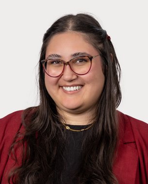 Eine lächelnde Frau mit langen, dunklen Haaren trägt eine rote Jacke und eine schwarze Bluse. Sie hat eine Brille und steht vor einem neutralen Hintergrund.