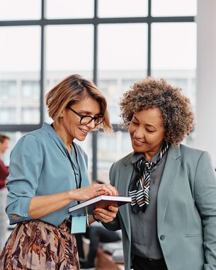 Zwei Frauen in formeller Kleidung stehen in einem modernen Raum und betrachten gemeinsam ein Buch. Im Hintergrund sind weitere Personen in einer Konferenz- oder Seminarumgebung zu sehen.