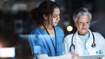 Zwei medizinische Fachkräfte, eine Krankenschwester und eine Ärztin, betrachten gemeinsam ein Tablet in einem klinischen Umfeld.