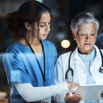 Zwei medizinische Fachkräfte, eine Krankenschwester und eine Ärztin, betrachten gemeinsam ein Tablet in einem klinischen Umfeld.