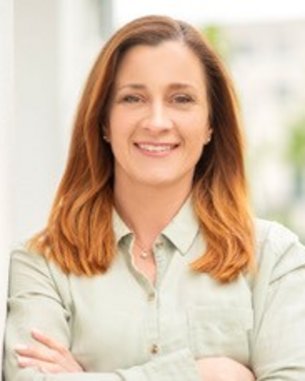 Eine Frau mit langen, braunen Haaren steht lächelnd mit verschränkten Armen vor einem unscharfen Hintergrund. Sie trägt ein helles Hemd und wirkt selbstbewusst.