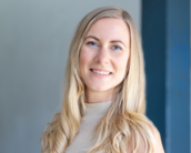 Eine lächelnde Frau mit langen, blonden Haaren trägt ein helles Oberteil. Der Hintergrund ist unscharf und zeigt eine Wand.