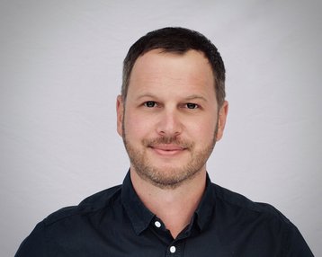 Ein Mann mit kurzem, dunklem Haar und einem leichten Bart lächelt in die Kamera. Er trägt ein dunkelblaues Hemd und hat einen neutralen Gesichtsausdruck vor einem hellen Hintergrund.