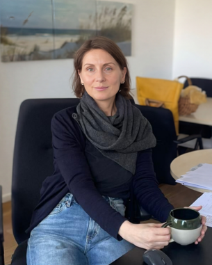 Frau mit einem schwarzen Schal sitzt an einem Schreibtisch und hält eine Tasse in der Hand, umgeben von Unterlagen und Büromaterial.