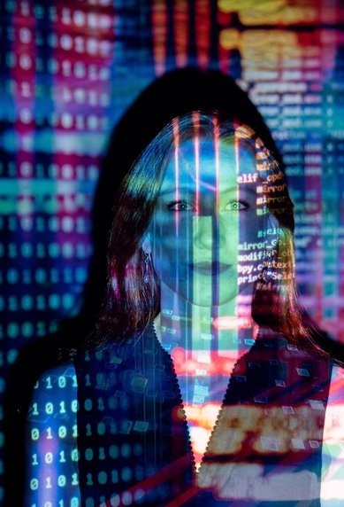 Eine Person steht vor einem Hintergrund aus bunten digitalen Codes und Datenströmen, die über ihr Gesicht projiziert werden. Die Darstellung vermittelt ein Gefühl von Technologie und Datenanalyse.