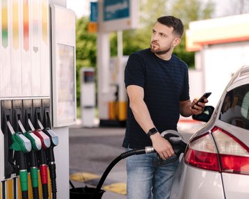 Ein Mann tankt sein Auto an einer Tankstelle und schaut auf sein Smartphone. Mehrere Zapfsäulen sind im Hintergrund sichtbar.