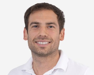 Ein Mann mit kurzen, braunen Haaren und einem freundlichen Lächeln trägt ein weißes Polohemd. Der Hintergrund ist neutral und hell.