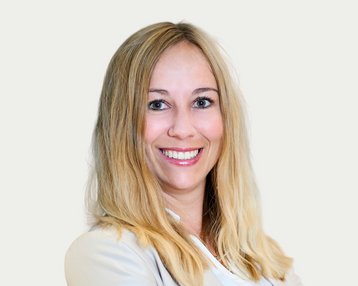 Eine lächelnde Frau mit langen, blonden Haaren trägt einen hellen Blazer und steht vor einem neutralen Hintergrund.