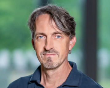 Ein Mann mit kurzen, grauen Haaren und einem Ohrring trägt ein dunkelblaues Polohemd. Er steht vor einem unscharfen Hintergrund mit grünen Elementen.