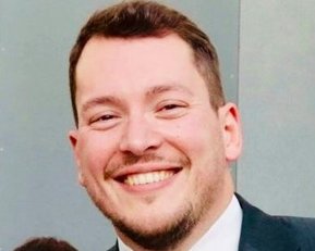 Ein Mann in einem Anzug lächelt freundlich in die Kamera, während er ein Schild hält.