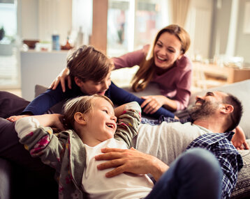 Eine Familie sitzt auf einem Sofa und lacht zusammen. Zwei Kinder spielen mit ihrem Vater, während die Mutter lächelnd zusieht.