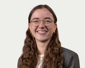 Eine lächelnde Frau mit langen, lockigen Haaren und Brille trägt einen Blazer. Der Hintergrund ist neutral und hell.