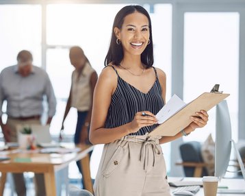 Eine Frau mit Clipboard und Notizen lächelt in einem modernen Büro, während Kollegen im Hintergrund an einem Tisch arbeiten.
