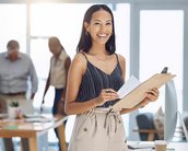 Eine Frau mit Clipboard und Notizen lächelt in einem modernen Büro, während Kollegen im Hintergrund an einem Tisch arbeiten.