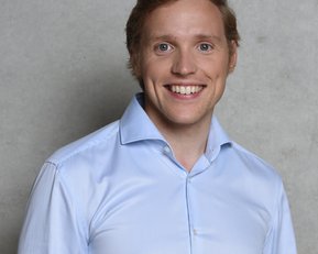 Ein junger Mann mit blonden Haaren und einem breiten Lächeln trägt ein hellblaues Hemd. Der Hintergrund ist neutral und grau.
