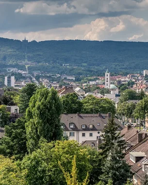 Blick auf eine Stadtlandschaft mit grünen Bäumen, Wohngebäuden und einer Kirche im Hintergrund, umgeben von Hügeln.