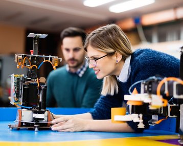Eine Frau mit Brille und ein Mann beobachten einen Roboter, während sie an einem Tisch in einer Werkstatt arbeiten. Der Roboter ist auf einem blauen Tisch platziert, umgeben von verschiedenen Werkzeugen und Geräten.