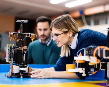 Eine Frau mit Brille und ein Mann beobachten einen Roboter, während sie an einem Tisch in einer Werkstatt arbeiten. Der Roboter ist auf einem blauen Tisch platziert, umgeben von verschiedenen Werkzeugen und Geräten.