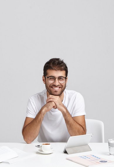 Ein junger Mann mit Brille sitzt an einem Tisch, lächelt und stützt sein Kinn auf die Hände. Vor ihm liegen Bücher, ein Notizbuch und eine Tasse. Ein Tablet steht auf dem Tisch.