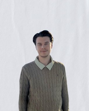 Junger Mann mit dunklen Haaren, der einen beigen Pullover mit Zopfmuster und einem hellen Kragen trägt. Er steht vor einem neutralen Hintergrund und schaut direkt in die Kamera.