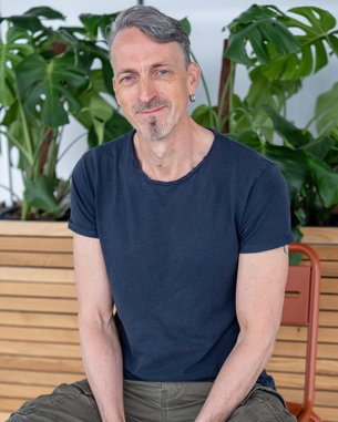 Ein Mann mit kurzen grauen Haaren und einem Ohrstecker sitzt auf einem Stuhl, lächelt und trägt ein schwarzes T-Shirt und grüne Hose. Pflanzen im Hintergrund.