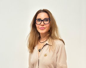 Eine Frau mit langen, lockigen Haaren und großen Brillen trägt ein beiges Oberteil und eine Halskette. Sie schaut direkt in die Kamera.