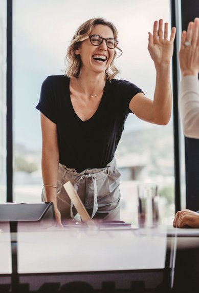 Eine Frau mit Brille steht an einem Tisch und gibt einer anderen Person ein High-Five. Beide lächeln, während sie in einem modernen Büro mit großen Fenstern sitzen.